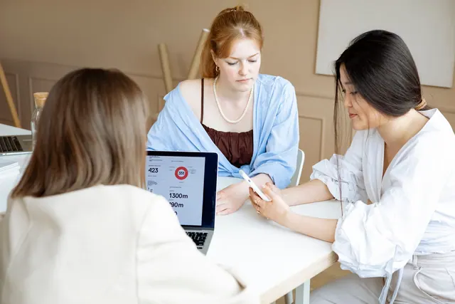 Three professional women discuss business strategy at the office table with laptops and documents. Grants for women comparison explained with examples